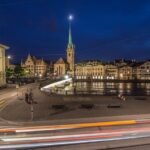 Nighttime cityscape along a river with illuminated historic buildings and church spires, and light trails from passing traffic in the foreground.