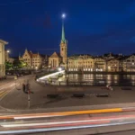 Nighttime view of Zurich's historic Old Town along the Limmat River, featuring illuminated churches, elegant waterfront buildings, and light trails from passing traffic.