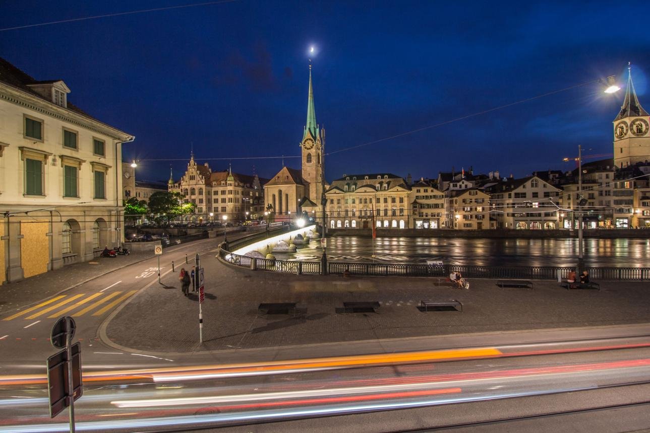 Nighttime cityscape along a river with illuminated historic buildings and church spires, and light trails from passing traffic in the foreground.
