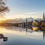Early morning sunlight reflecting across Lake Zurich with historic church spires, waterfront buildings, and the Swiss Alps in the background.