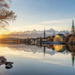 Early morning sunlight reflecting across Lake Zurich with historic church spires, waterfront buildings, and the Swiss Alps in the background.